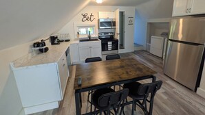 Eine kompakte Küche mit weißen Schränken, einem Edelstahlkühlschrank, einer Mikrowelle und einer Kaffeemaschine. Ein Holztisch mit vier Stühlen steht daneben.