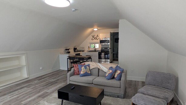 Ein Loft-Wohnbereich mit grauem Sofa, grauem Sessel und einem schwarzen Couchtisch. Im Hintergrund ist eine Küche mit weißen Schränken und einer Mikrowelle erkennbar.