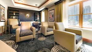 A hotel room with a seating area featuring two armchairs and a small table, a bookshelf, and a large window with a city view.