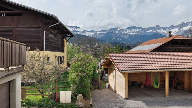 A rustic building with a wooden exterior and a red-tiled roof, surrounded by a gravel driveway and a small garden with flowering plants. - Bed and Breakfast 'La Ferme Du Mont Blanc - Gouter' with Mountain View and Wi-Fi (Combloux)