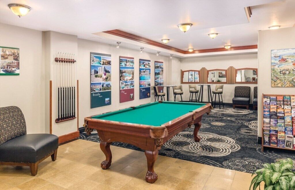 A pool table with a green felt surface, surrounded by a carpeted area with a patterned design. There are several framed pictures on the wall, a seating area with chairs, and a small table with a lamp.