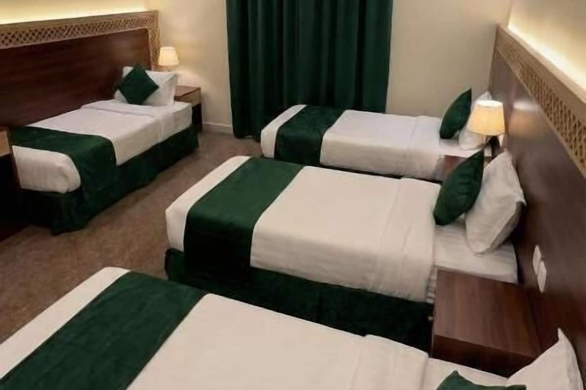 Et hotelværelse med tre enkeltsenge, hver med et grønt og hvidt sengetæppe og matchende puder. Værelset har en træsenggavl, et stort vindue med gardin i grøn farve og en loftlampe.
