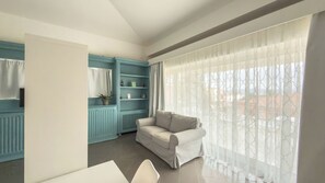 Ein modernes Wohnzimmer mit einer hellblauen Einbauschränkenwand, einem beigen Sofa und einem großen Fenster mit transparenten Vorhängen.