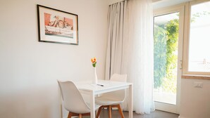 Een eethoek met een witte tafel en stoelen, een vaas met bloemen, een ingelijste foto aan de muur en uitzicht op groen door het raam.