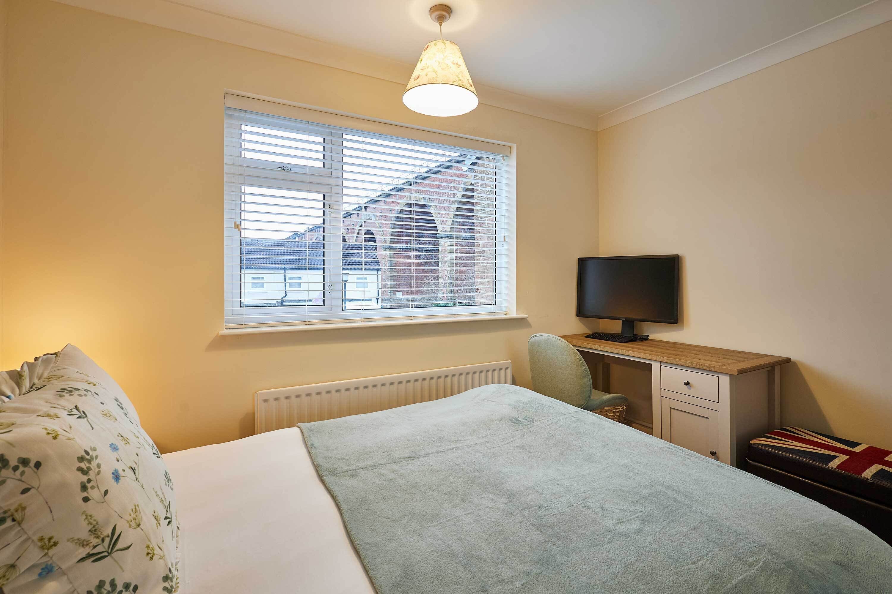 A bedroom with a bed, a desk, a chair, a television, and a window with blinds.