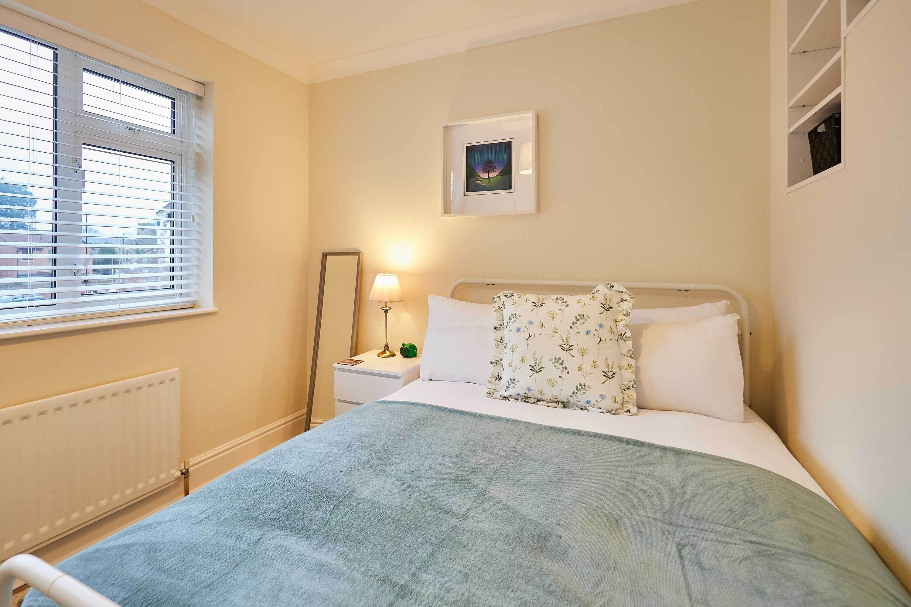 Une chambre à coucher avec un lit, une table de chevet avec une lampe, une fenêtre avec des stores, un radiateur et un tableau encadré au mur.