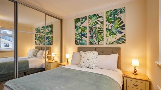 A bedroom with a bed, two bedside tables, a mirror, and tropical-themed wall art. - Host & Stay | Bridge Court (Yarm)