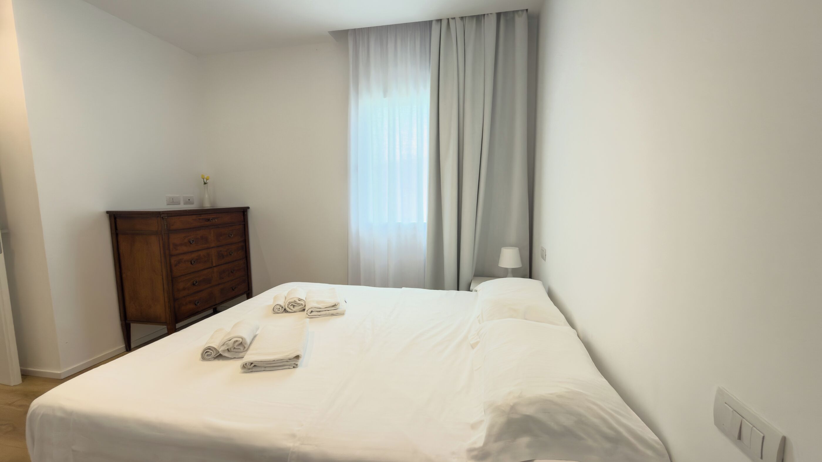 Un lit bien fait, avec du linge de lit blanc et deux serviettes enroulées, une commode en bois et une fenêtre ornée de rideaux légers.