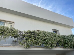 Un bâtiment moderne à la façade blanche, doté d’un balcon orné de plantes grimpantes à fleurs blanches, et surplombant un ciel d’un bleu limpide.