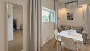 A modern living room with a sofa, a dining table, and chairs. There is a window with curtains, an air conditioning unit, and a small decorative item on the windowsill. - Villa Ada-Orazio - One Bedroom Apartment, Sleeps 2 (Olbia)