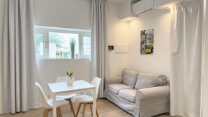 A modern, minimalist room with a white table and chairs, a beige sofa, and a window with curtains. - Villa Ada-Orazio - One Bedroom Apartment, Sleeps 2 (Olbia)