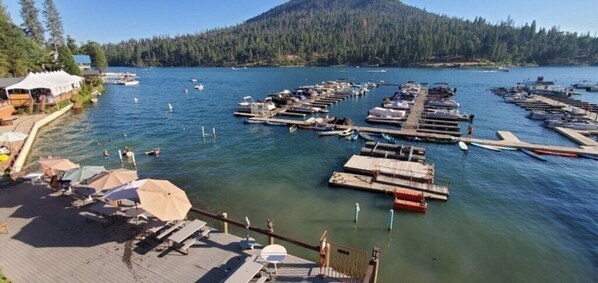 A marina with boats docked, a dock with sun loungers, and a forested hill in the background.