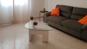 En stue med et glasbord, en sofa med orange puder, et lille sofabord med en plante og et blomsterbillede i ramme på væggen.