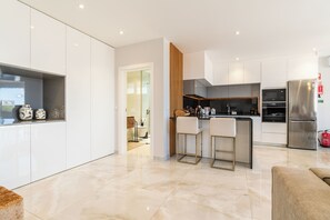 A modern kitchen with white cabinets, a stainless steel refrigerator, and a bar area with two stools. - El Dourado House | Ocean View (Caniço)