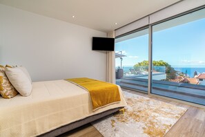 A bedroom with a bed, a large window, and a TV on the wall. - El Dourado House | Ocean View (Caniço)