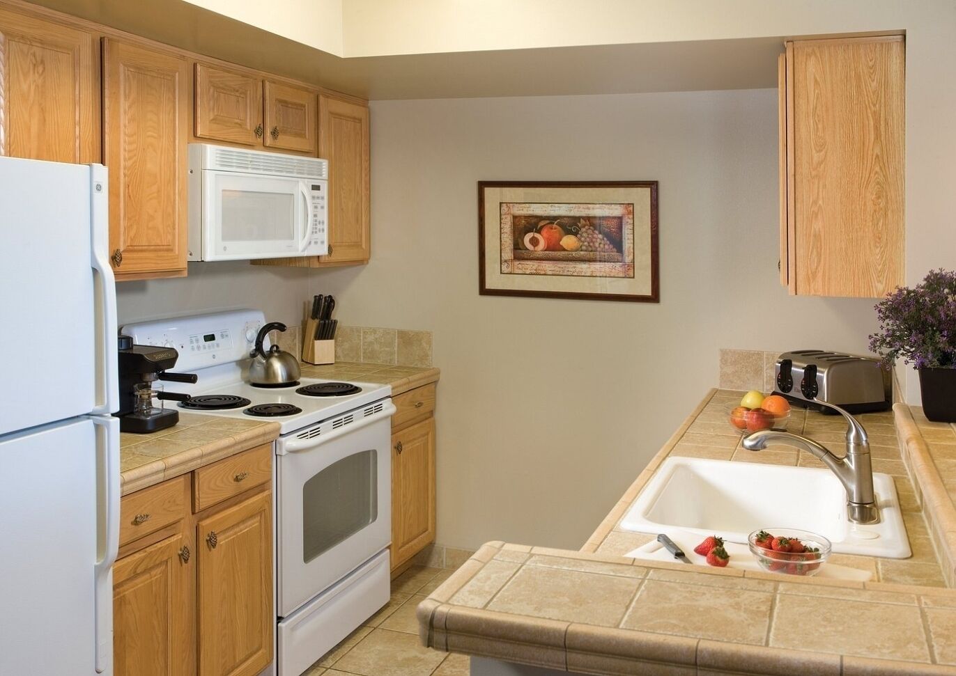 A kitchen with wooden cabinets, a white stove, a microwave, a kettle, a toaster, and a sink with fruit.