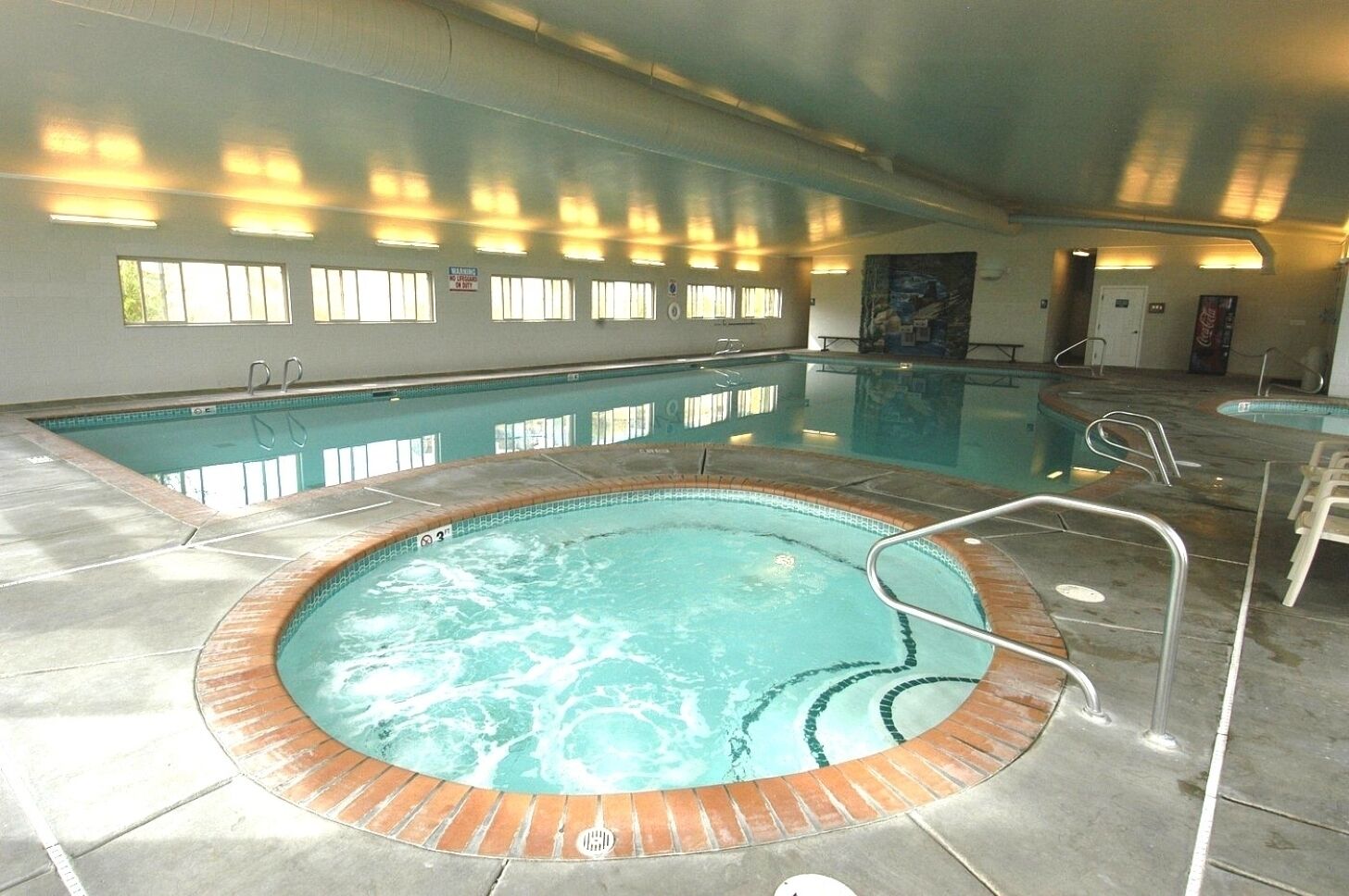 An indoor swimming pool with a hot tub, tiled walls, and a windowed wall.
