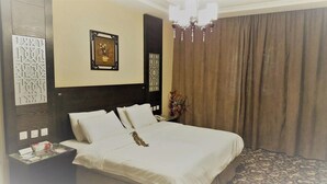A neatly made bed with white linens, a nightstand with a phone and papers, a framed picture above the bed, a chandelier, and a curtain on the window.