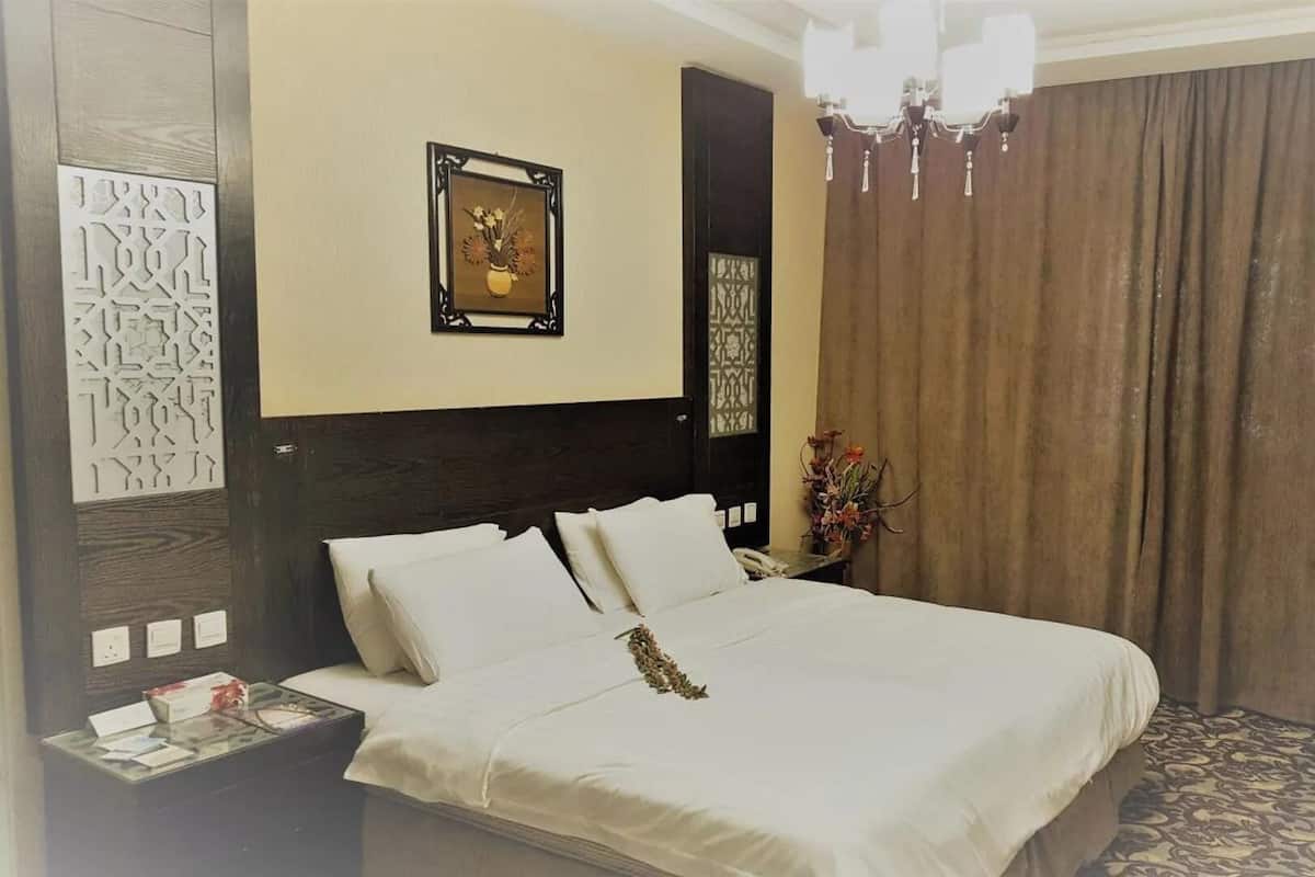 A neatly made bed with white linens, a nightstand with a phone and papers, a framed picture above the bed, a chandelier, and a curtain on the window.