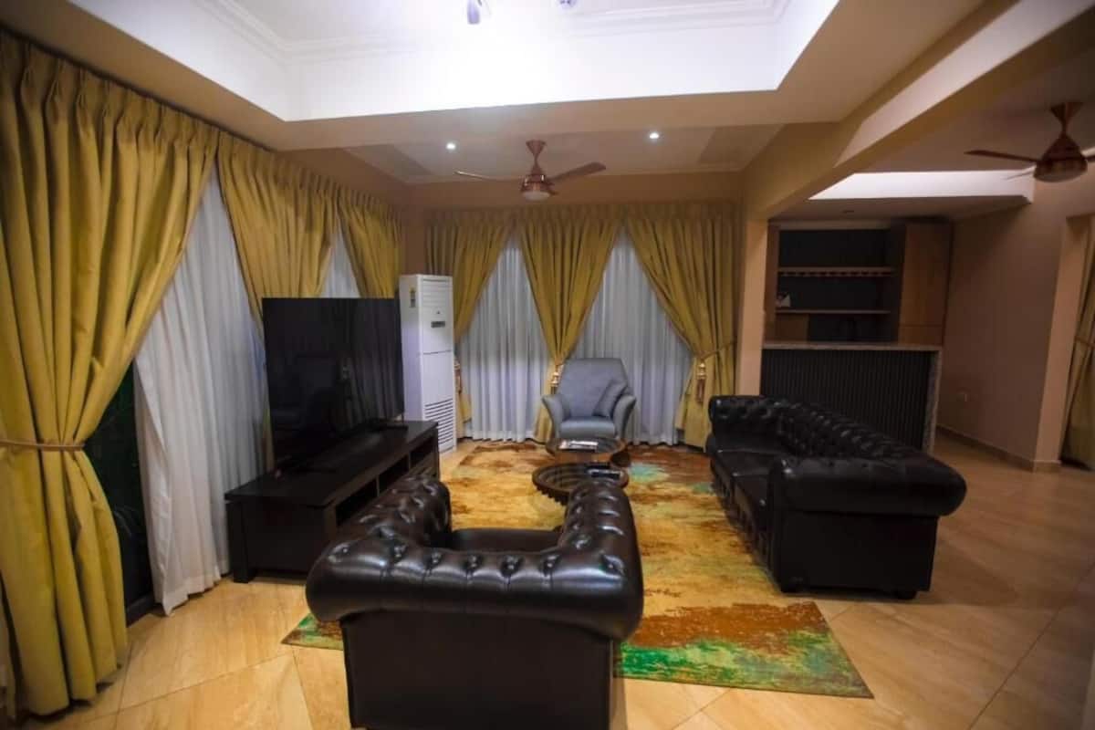 Una sala de estar con un sofá de cuero negro, un sillón gris y un reposapiés negro. Hay un televisor de pantalla plana sobre un mueble bajo, un ventilador de techo y un amplio ventanal con cortinas.