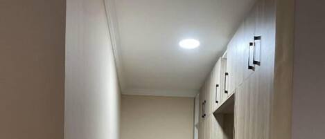 A narrow hallway with light-colored walls and a tiled floor. There is a black metal bench in the center and wooden cabinets on the right.