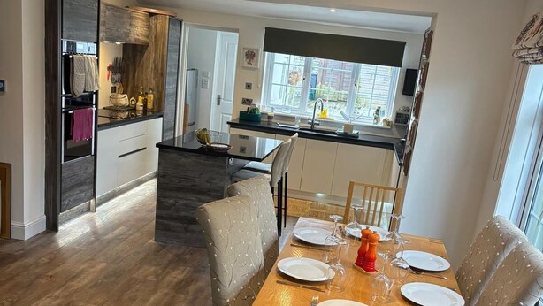 Un comedor con una mesa de madera dispuesta para cuatro, una cocina al fondo y vistas al exterior a través de una ventana.