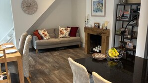 Una sala moderna con comedor, que presenta un comedor de madera, dos taburetes de bar, un sofá gris con cojines florales, una chimenea y una estantería.