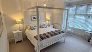 Una cama con dosel sobre una base de cuatro postes, mesitas de noche con lámparas, un radiador y una ventana con persianas.