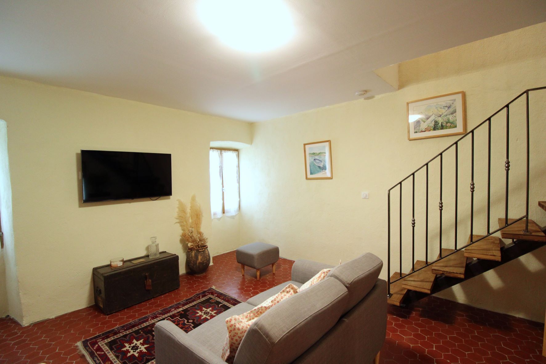 A living room with a sofa, a TV, a small table, a vase with dried plants, a rug, a staircase, and a window.