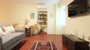 Ein Wohnzimmer mit grauem Sofa, einem kleinen Esstisch, einem gemusterten Teppich, einem Bücherregal und einem an der Wand montierten Fernseher.