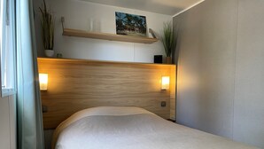 A neatly made bed in a hotel room with a wooden headboard, bedside lamps, and a small shelf with decorative items. - Bungalow in Lège-Cap-Ferret between pine forest, lake and ocean (Lège-Cap-Ferret)