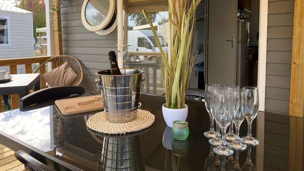 A wooden deck with a table set for a meal, a bottle of champagne, and a vase with plants. - Bungalow in Lège-Cap-Ferret between pine forest, lake and ocean (Lège-Cap-Ferret)