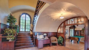 A grand staircase leading to a second floor, a billiards table, and a cozy seating area with a lamp and books.