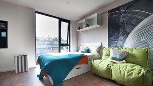 A modern bedroom with a large window, a green sofa, a bed with a blue blanket, and a cityscape view. - Student Only Gateway Apartments (Edinburgh)