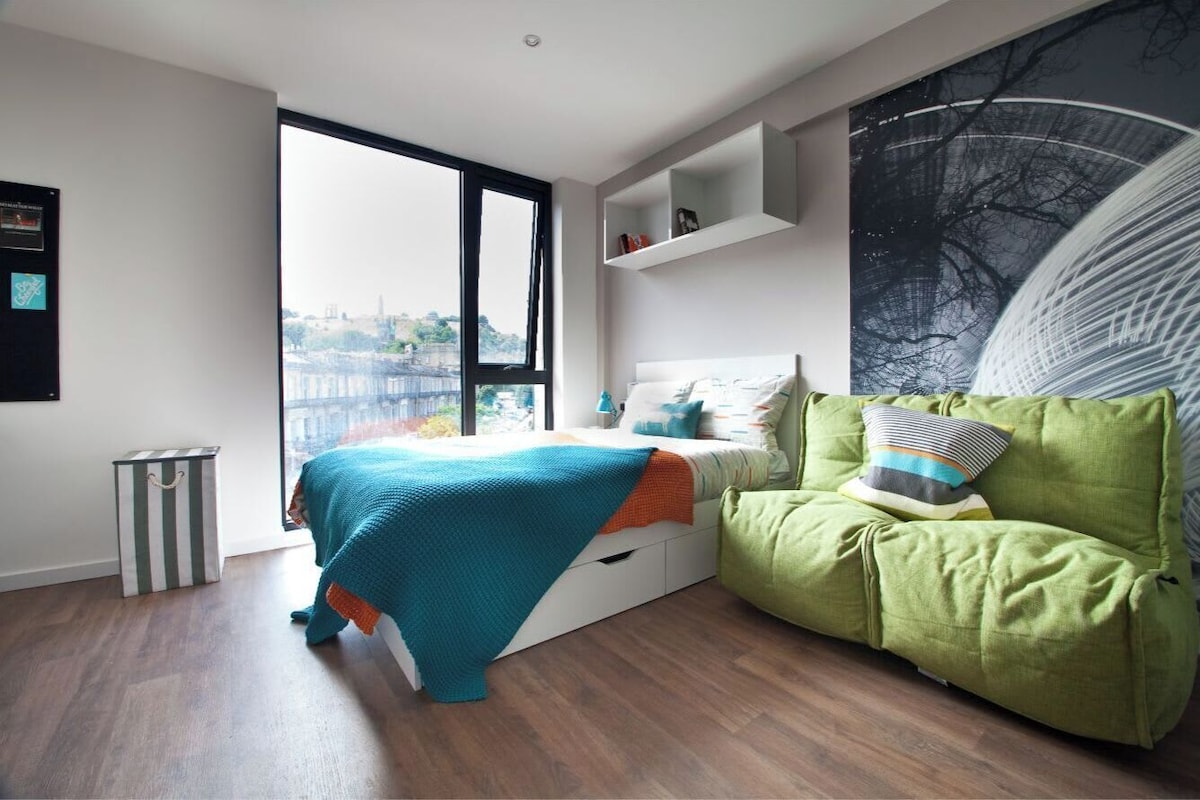 A modern bedroom with a large window, a green sofa, a bed with a blue blanket, and a cityscape view.