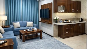 Una habitación de hotel moderna con un sofá azul, una mesa de centro de madera, un televisor de pantalla plana, una kitchenette con un lavamanos y armarios, y un equipo de aire acondicionado.