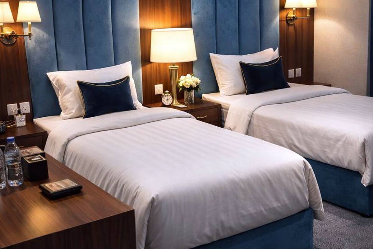 A hotel room with two beds, each with white linens and dark blue headboards. There are bedside tables with lamps, a clock, and a vase with flowers.