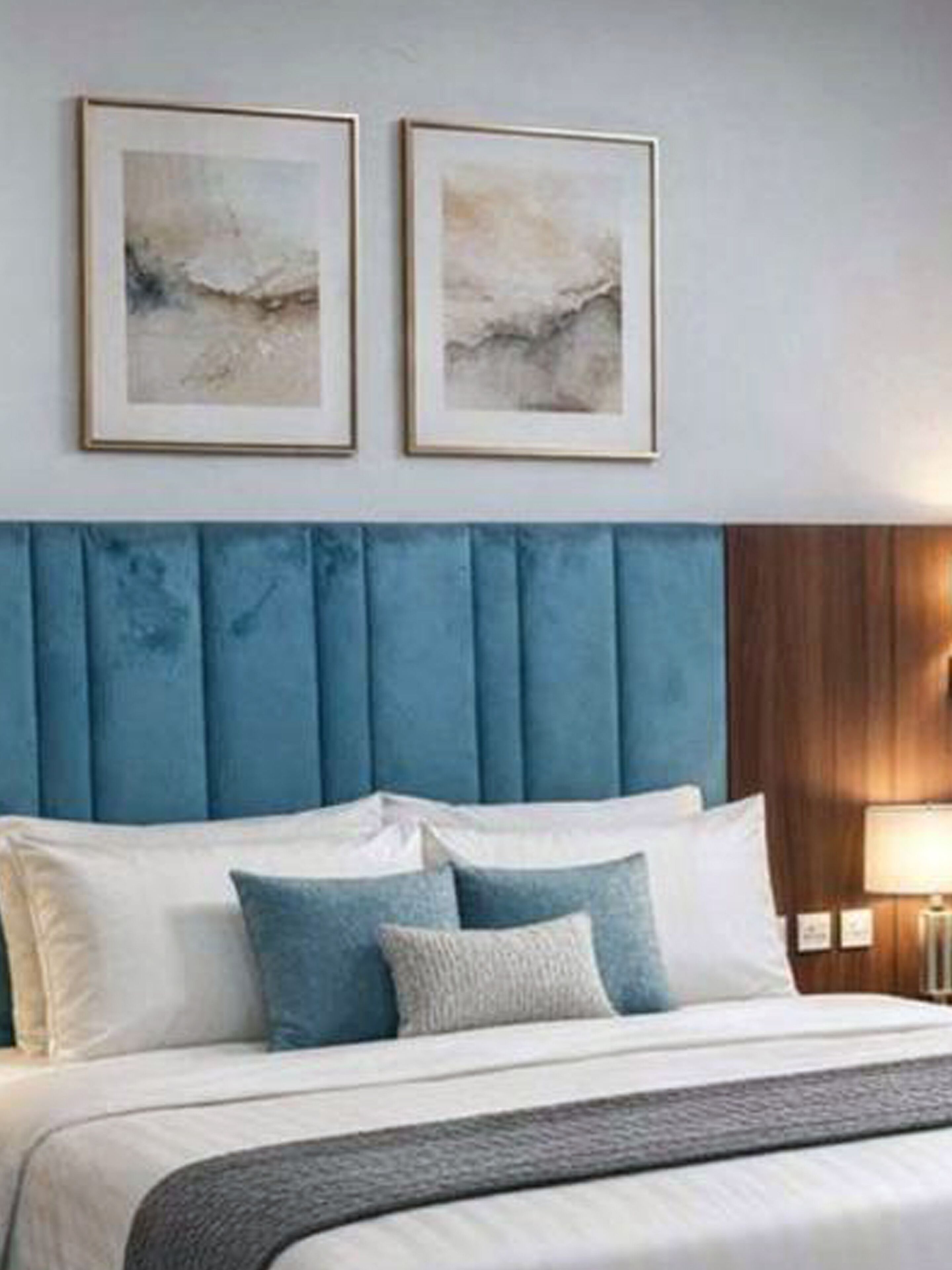 A bedroom with a blue upholstered headboard, a bed with white and blue bedding, and two framed abstract paintings on the wall.