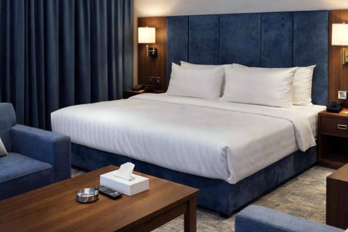 A hotel room with a large bed, a bedside table with a lamp, a chair, and a small table with a tissue box and remote control.