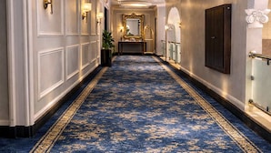 Un pasillo con alfombra azul, apliques de pared y un espejo.
