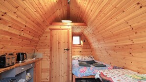 A cozy attic bedroom with wooden walls, a small window, a bed with floral bedding, a kitchenette with a kettle, and a patterned floor.