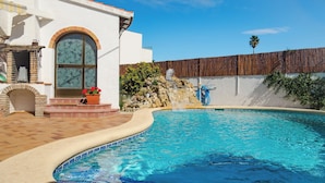 Une piscine agrémentée d’une cascade en pierre, une terrasse en briques et un bâtiment blanc doté d’une grande fenêtre en arc de cercle.
