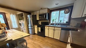 Une cuisine avec un plancher en bois, des appareils électroménagers en acier inoxydable, un coin repas avec une table et des chaises, et une fenêtre donnant sur les arbres.