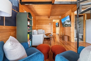 A cozy cabin interior with a sofa, ottomans, a bookshelf, and a television. - Lake Granby View Cabin | Sleeps 8, Hot Tub & Firepits (Granby)