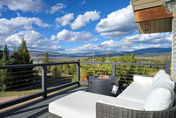 Un balcón con un sofá blanco para exteriores, una chimenea y vistas a montañas y un lago.