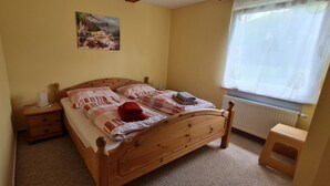A bedroom with a wooden bed, a nightstand, a small stool, a radiator, and a window with a curtain. - Comfortably furnished vacation home with fireplace (Ilsenburg (Harz))
