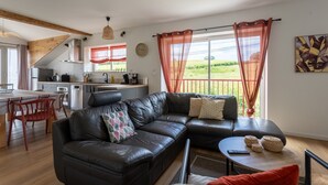 A modern living room with a black leather sofa, a dining area with red chairs, and a kitchen in the background. - Cottage in the Basque Mountains with View, Wi-Fi and Air Conditioning (Ossès)