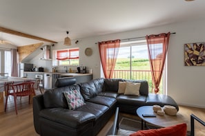 A modern living room with a black leather sofa, a dining area with red chairs, and a kitchen in the background. - Cottage in the Basque Mountains with View, Wi-Fi and Air Conditioning (Ossès)