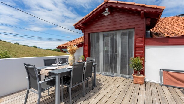 A wooden deck with a table and chairs, a red building with a tiled roof, and a potted plant. - Cottage in the Basque Mountains with View, Wi-Fi and Air Conditioning (Ossès)