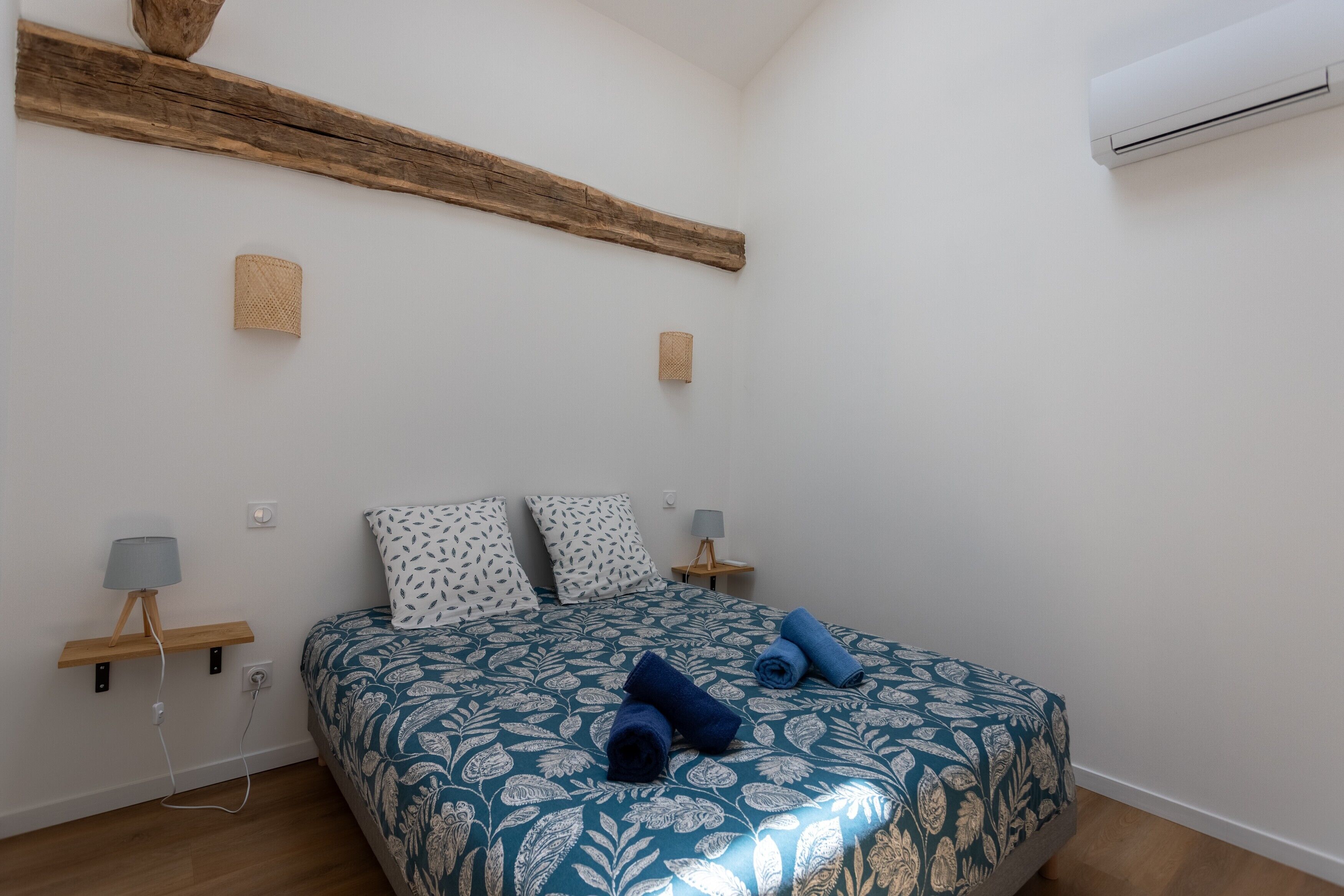 Un dormitorio con una cama, dos mesitas de noche, una lámpara, aire acondicionado instalado en la pared y una viga de madera.